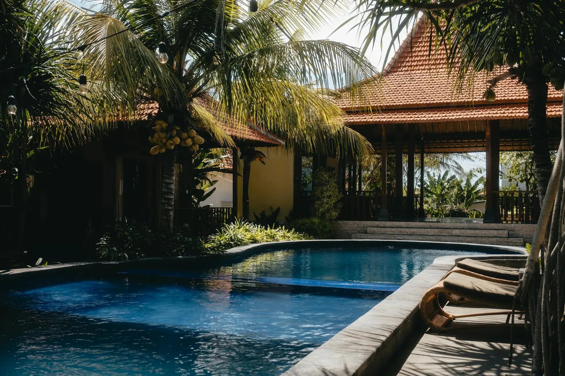 A luxurious tropical villa featuring a deep blue swimming pool surrounded by lush palm trees and traditional orange-tiled pavilions under bright sunlight.